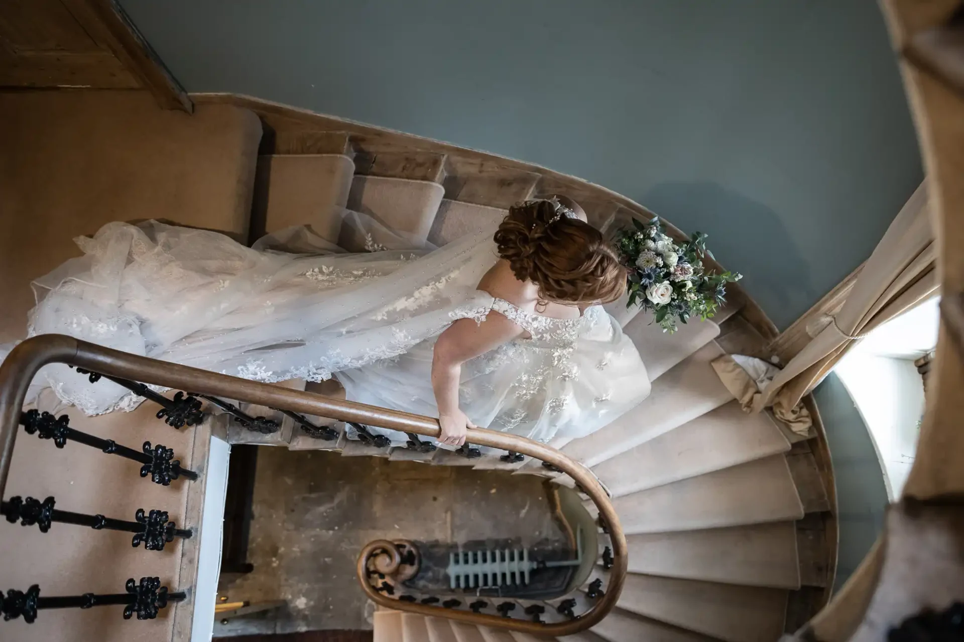 Bride in a white dress descends a spiral staircase, holding a bouquet, viewed from above.