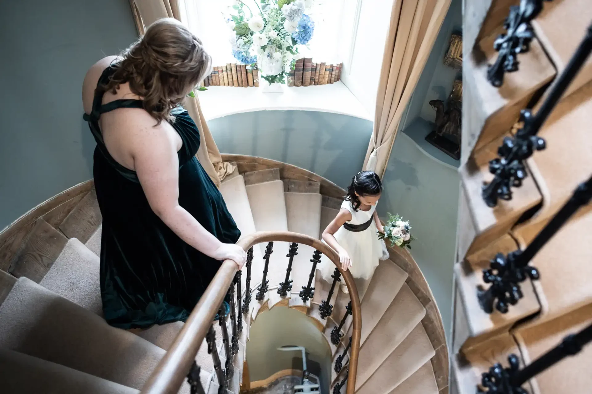 Woman in a green dress and girl in a white dress holding flowers walk down a spiral staircase beside a window with books and floral decor.