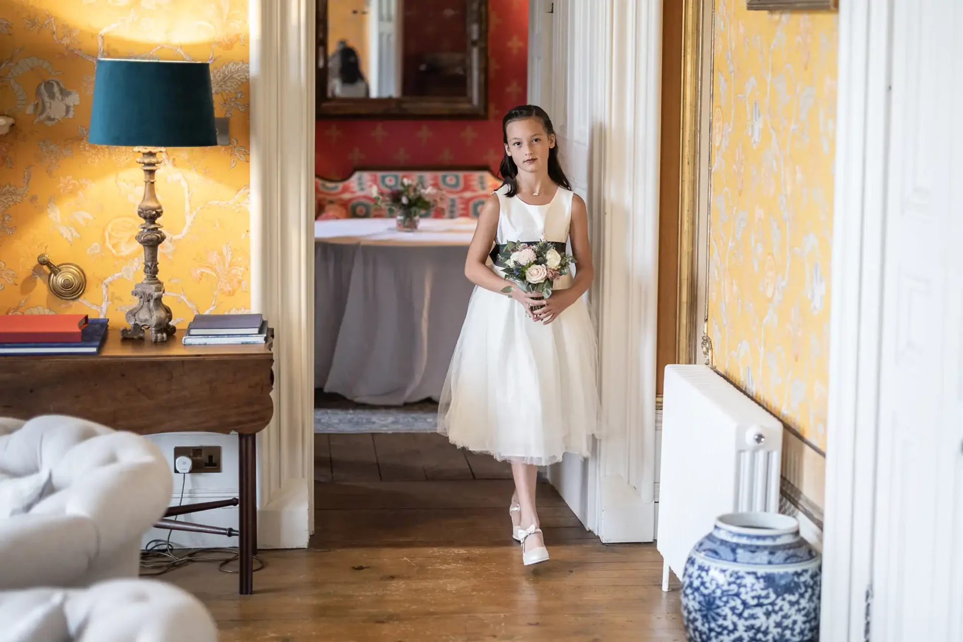 Young girl in a white dress holding a bouquet stands in a doorway, surrounded by yellow floral wallpaper and wooden flooring.