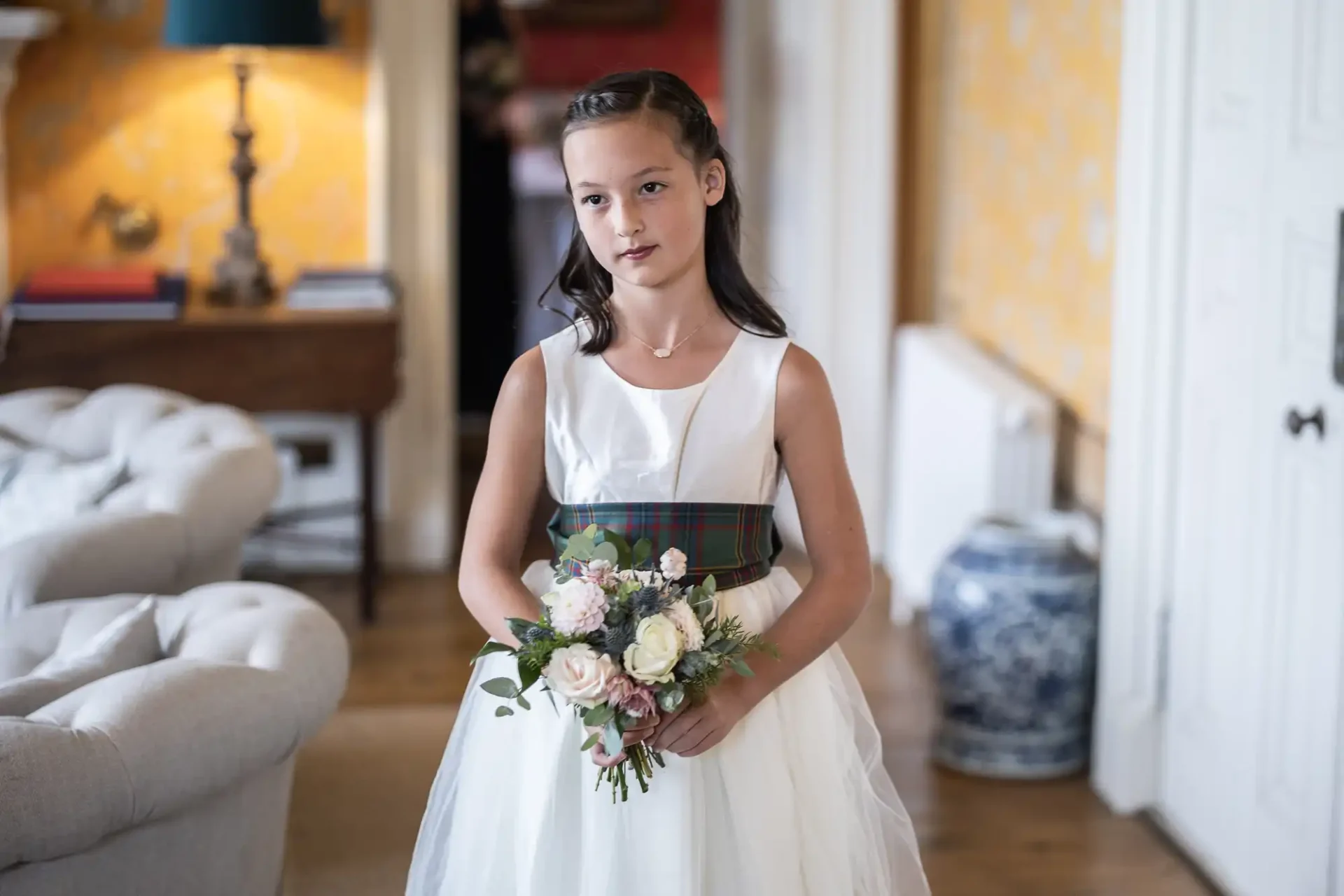 A young girl in a white dress holds a bouquet of flowers, standing indoors. A beige sofa, a lamp, and a decorative vase are visible in the background.