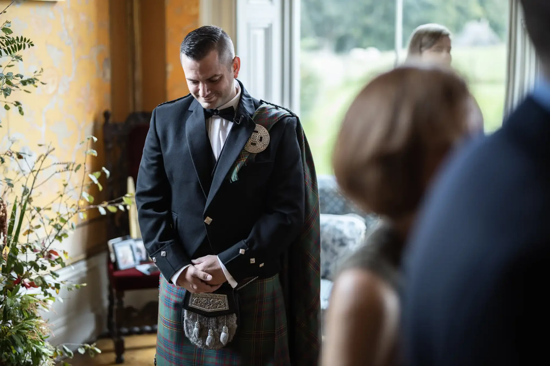 A man in formal attire, including a kilt, stands indoors with his head bowed and hands clasped. A few other people are partially visible in the room.