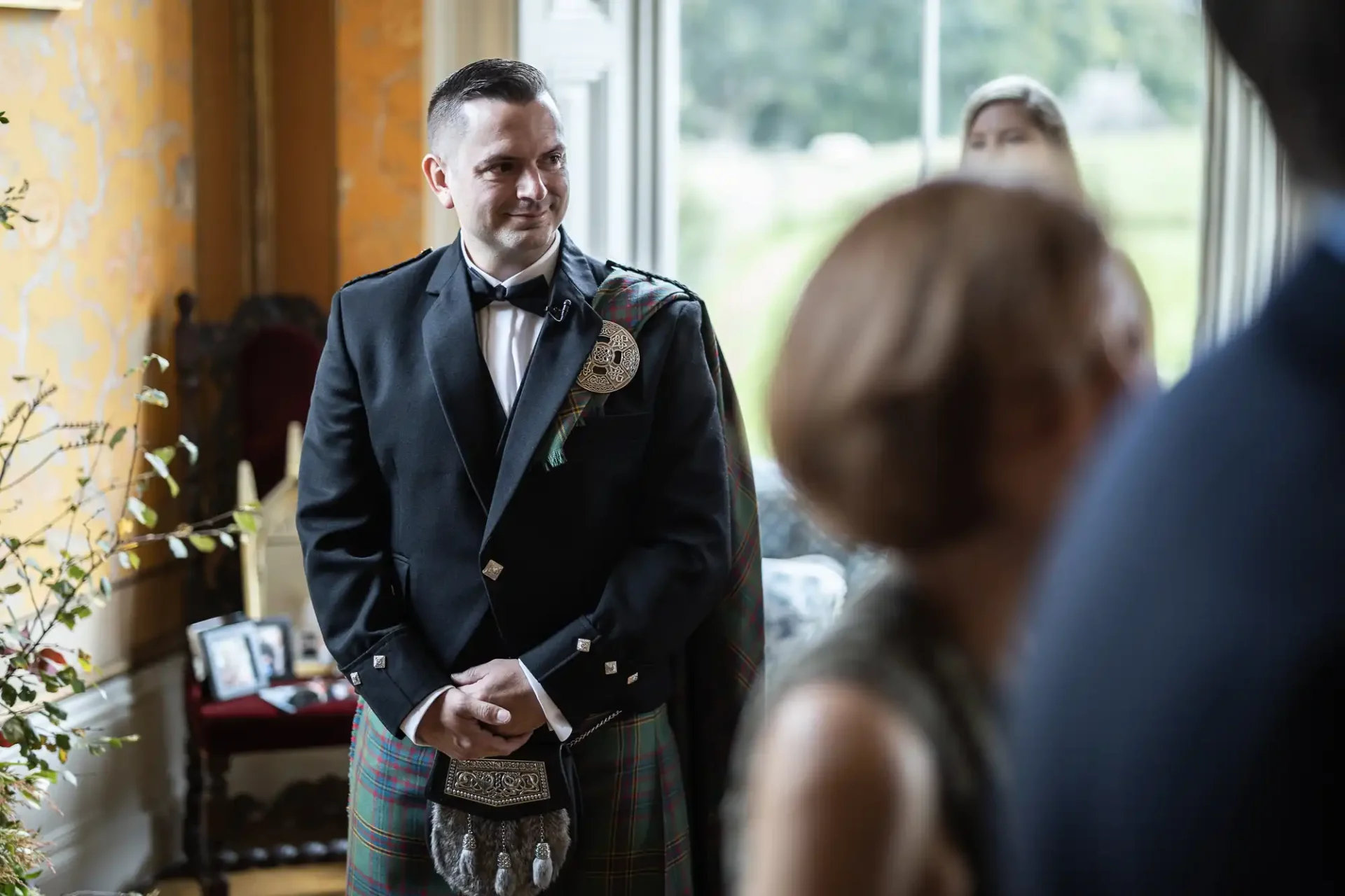 A man in formal Scottish attire stands indoors near a window, smiling. Other people are blurred in the foreground.