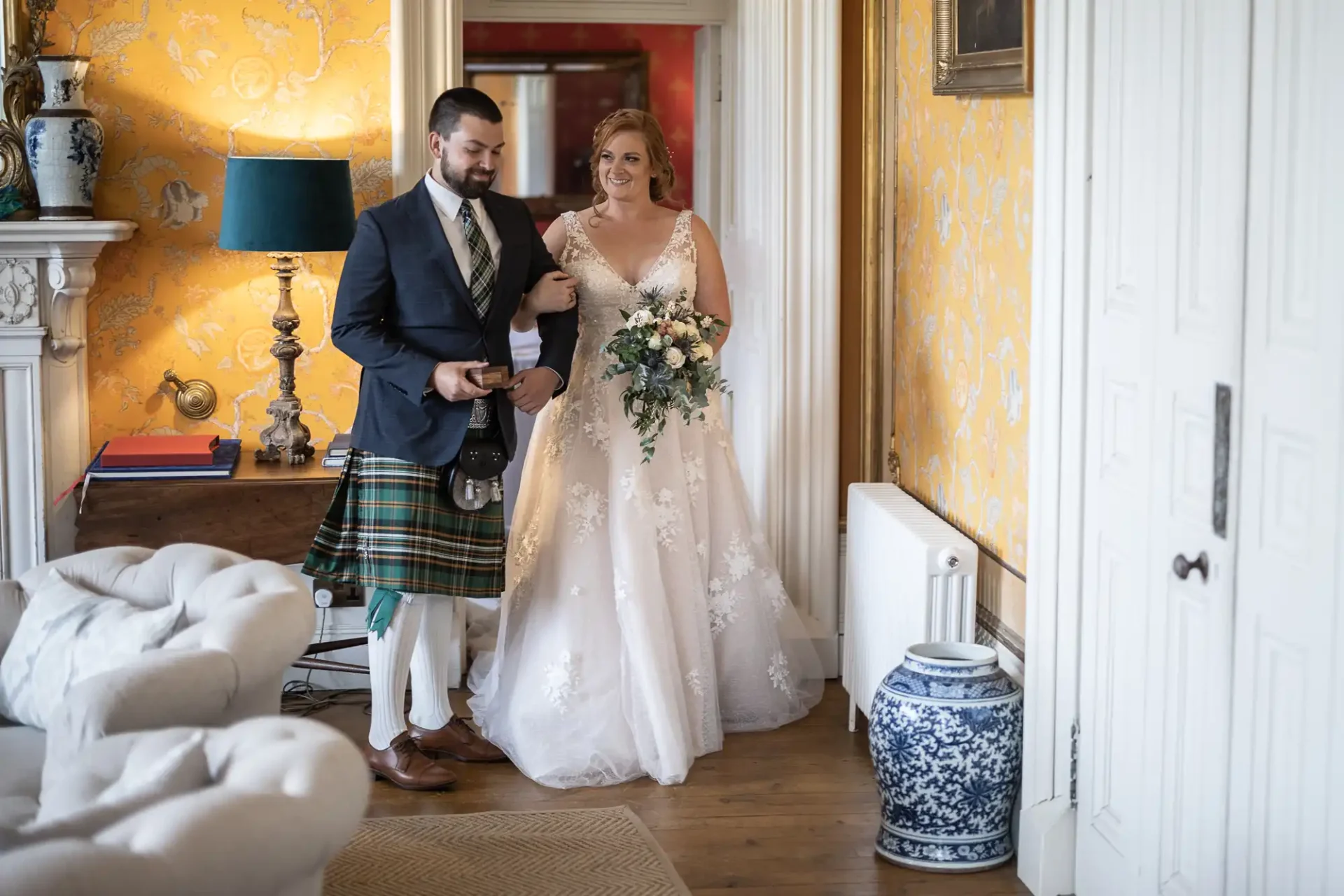 Bride in a white gown holding a bouquet and a man in a kilt walking together in a room with yellow patterned walls and ornate decor.