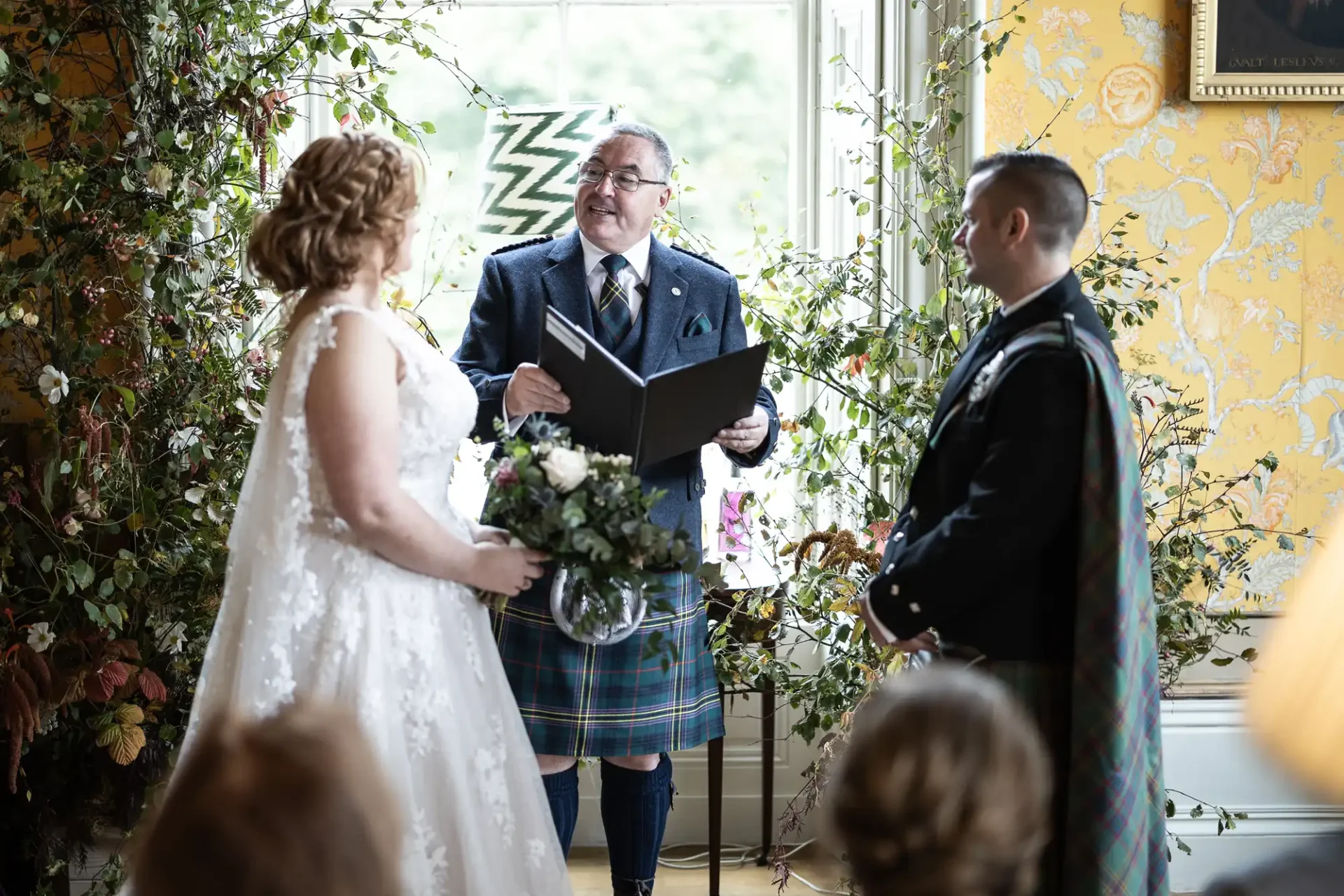 A wedding ceremony with a couple and an officiant wearing tartan attire. The bride holds a bouquet, and they stand before a window adorned with foliage.