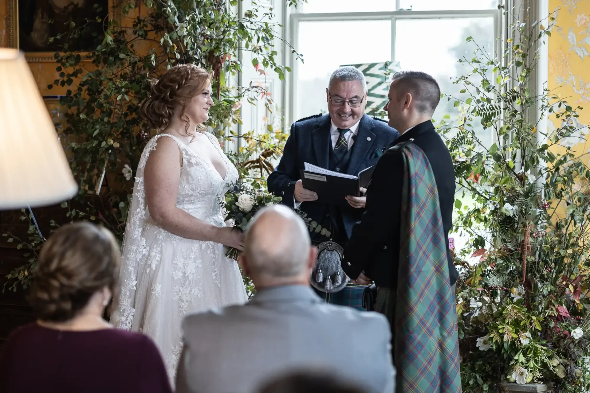 A couple in wedding attire stands before an officiant in a decorated room, exchanging vows. The groom wears a kilt. Guests are seated in the foreground.