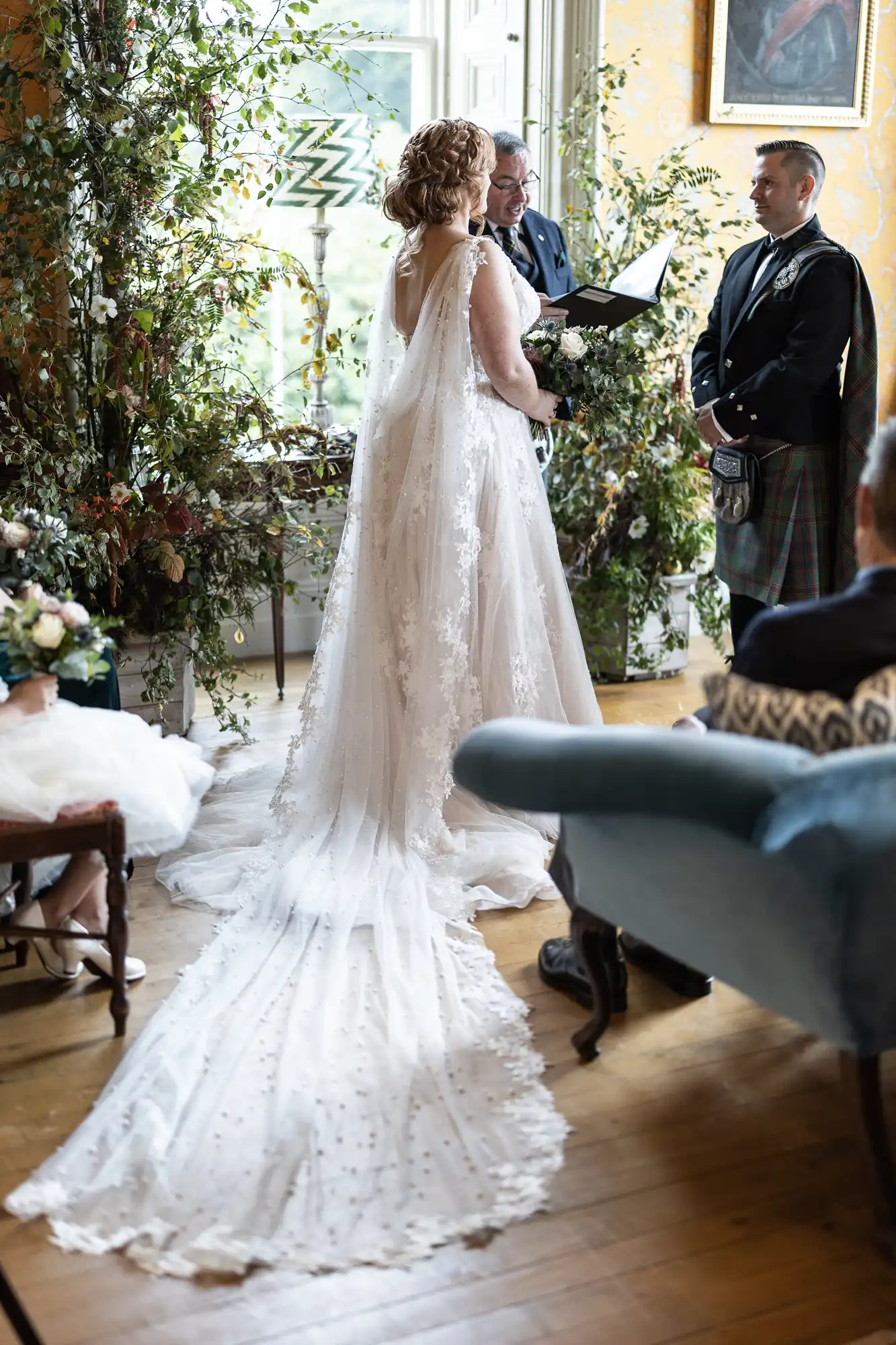 Bride in a white gown with a long train stands with a groom in a kilt during a wedding ceremony in a decorated room.