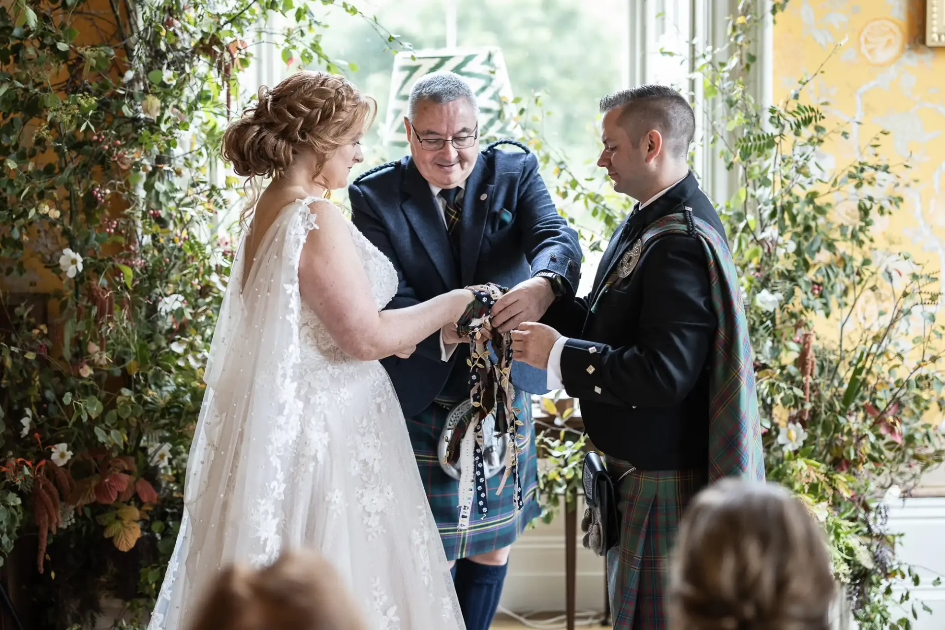 A couple in wedding attire participates in a handfasting ceremony officiated by a man in a kilt, surrounded by greenery and floral decor.