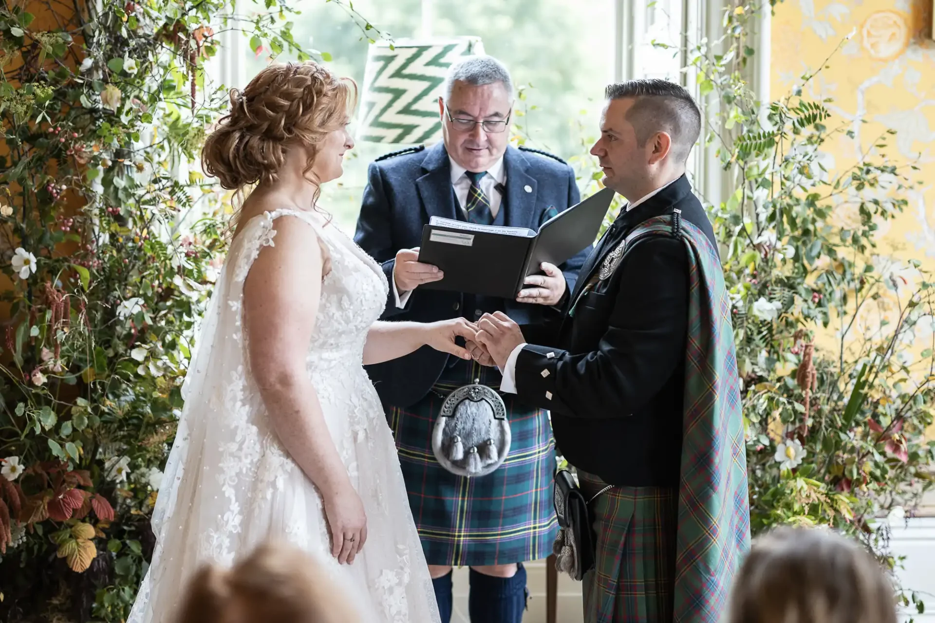 A couple exchanges vows during a wedding ceremony indoors. The groom and officiant wear kilts, while the bride is in a white gown. Floral decorations surround the window in the background.