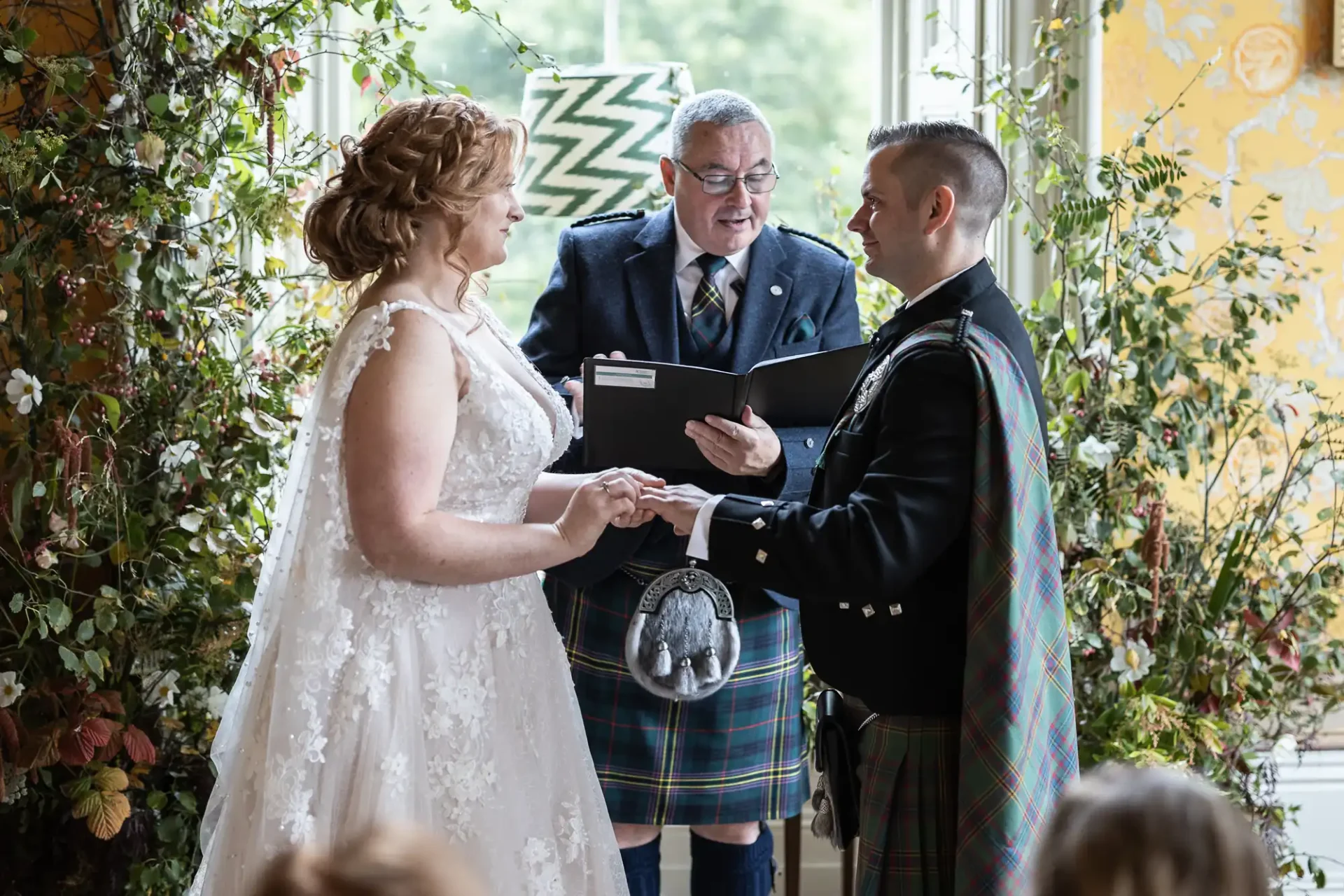 A couple in formal attire exchanges rings during a wedding ceremony, with an officiant standing between them. The setting includes colorful floral decorations and a large window.