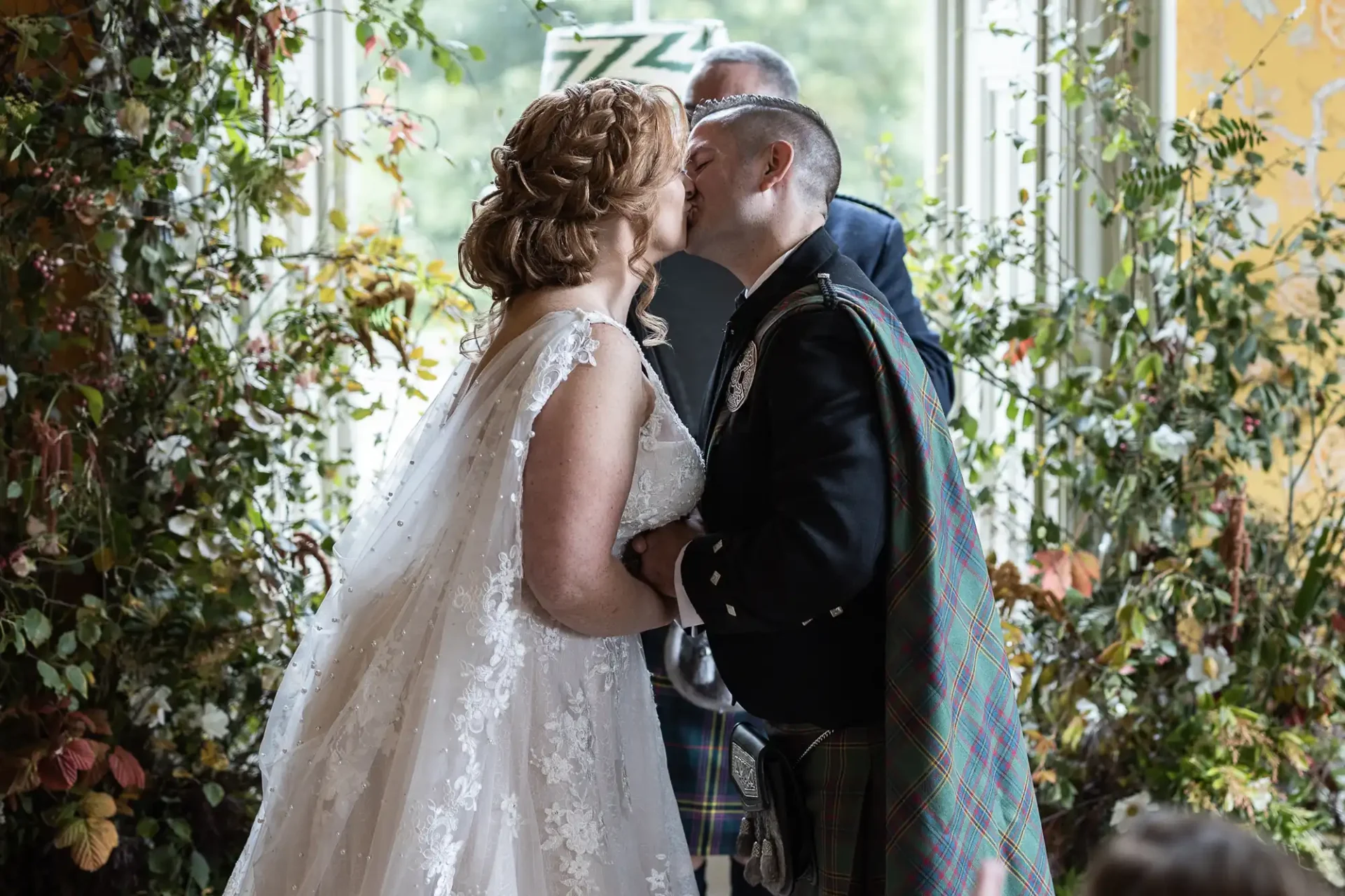 A couple kisses at their wedding ceremony. The bride is in a white gown, and the groom wears a traditional kilt. The setting is decorated with greenery and flowers.