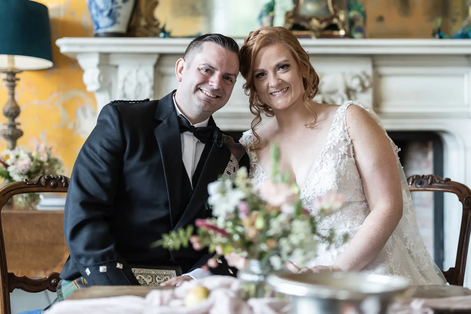 A man in a kilt and a woman in a white dress sit closely, smiling, in front of a decorated fireplace with a floral centerpiece on the table.