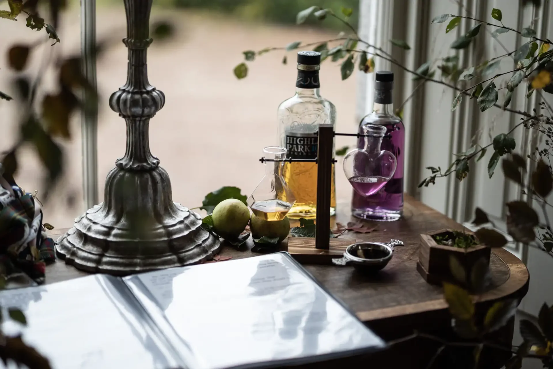 Bottles of alcohol and a green apple on a wooden table beside a window, with a large metal candle holder and scattered leaves.