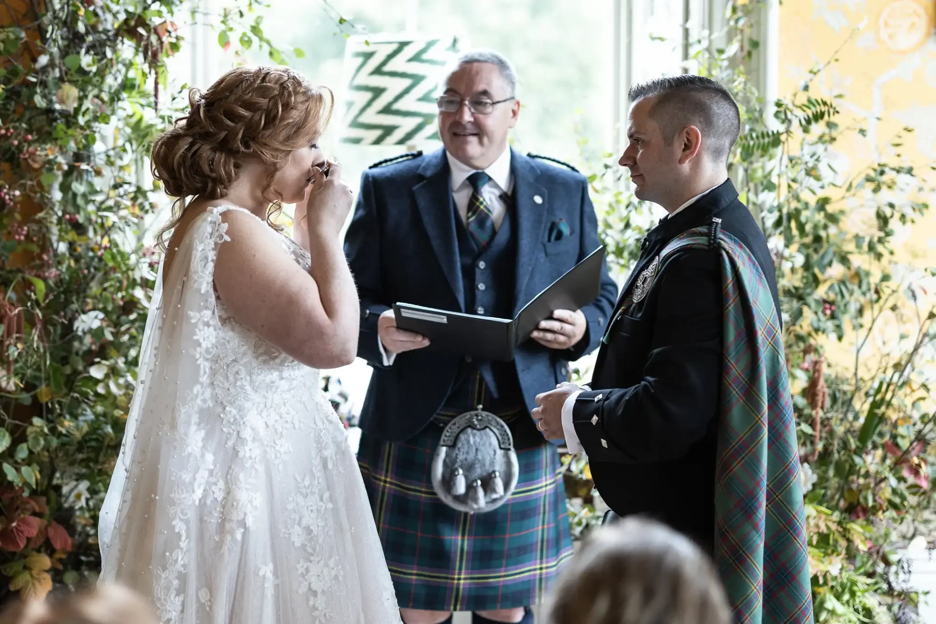 Bride and groom in kilts exchange vows in a floral setting with an officiant holding a book. Bride wipes tears, groom smiles.