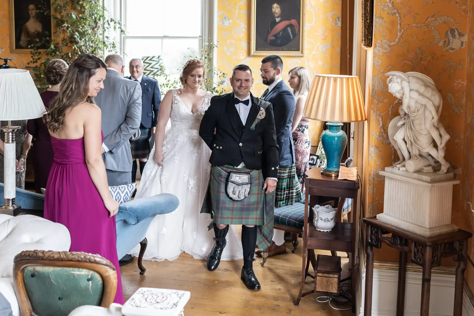 A wedding scene indoors with people in formal attire. A bride in a white gown smiles, and a groom in a kilt stands beside her. Others in dresses and kilts are around them in a decorated room.