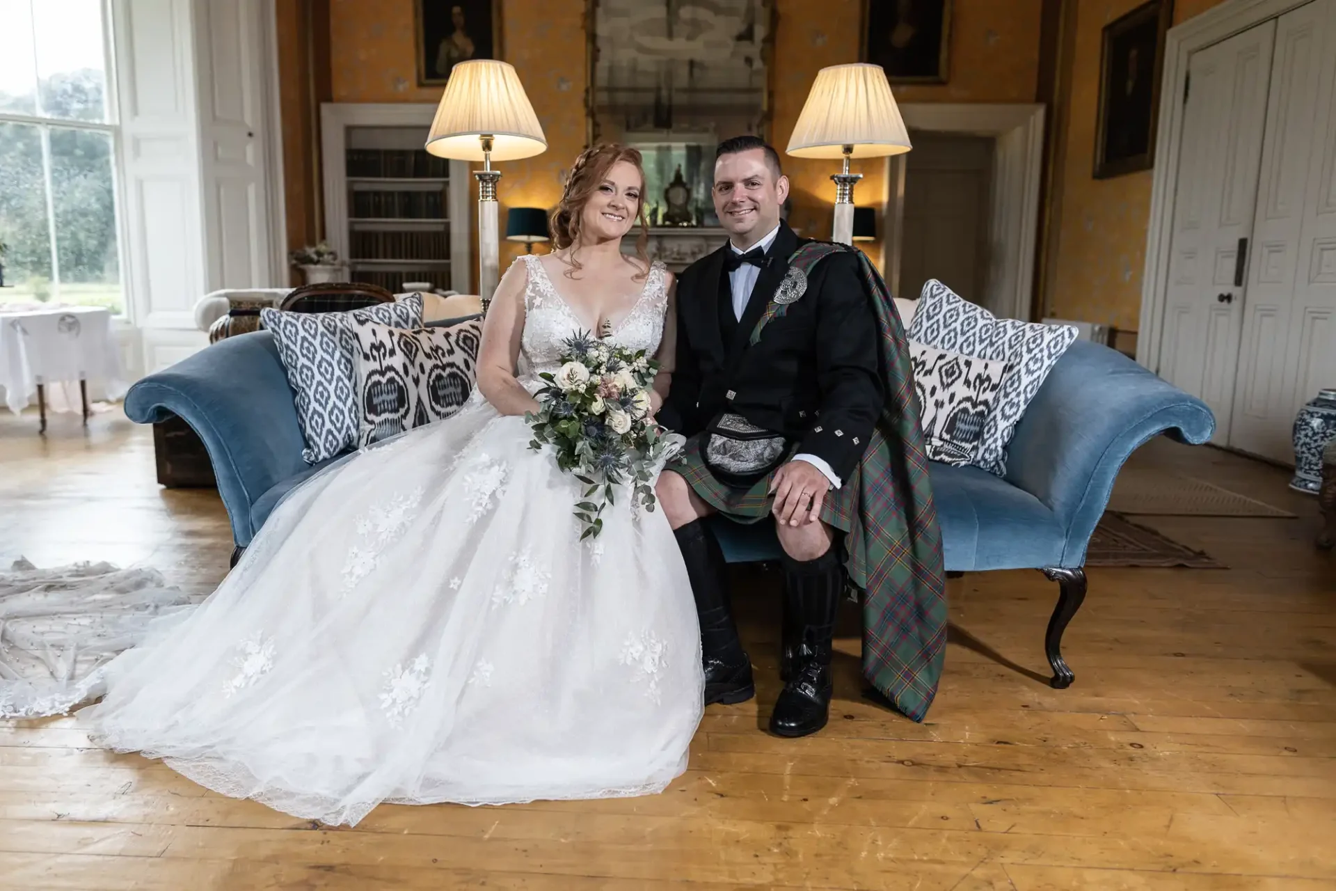 A couple in wedding attire sits on a blue couch in a room with wooden floors and lamps. The bride holds a bouquet, and the groom wears a kilt.