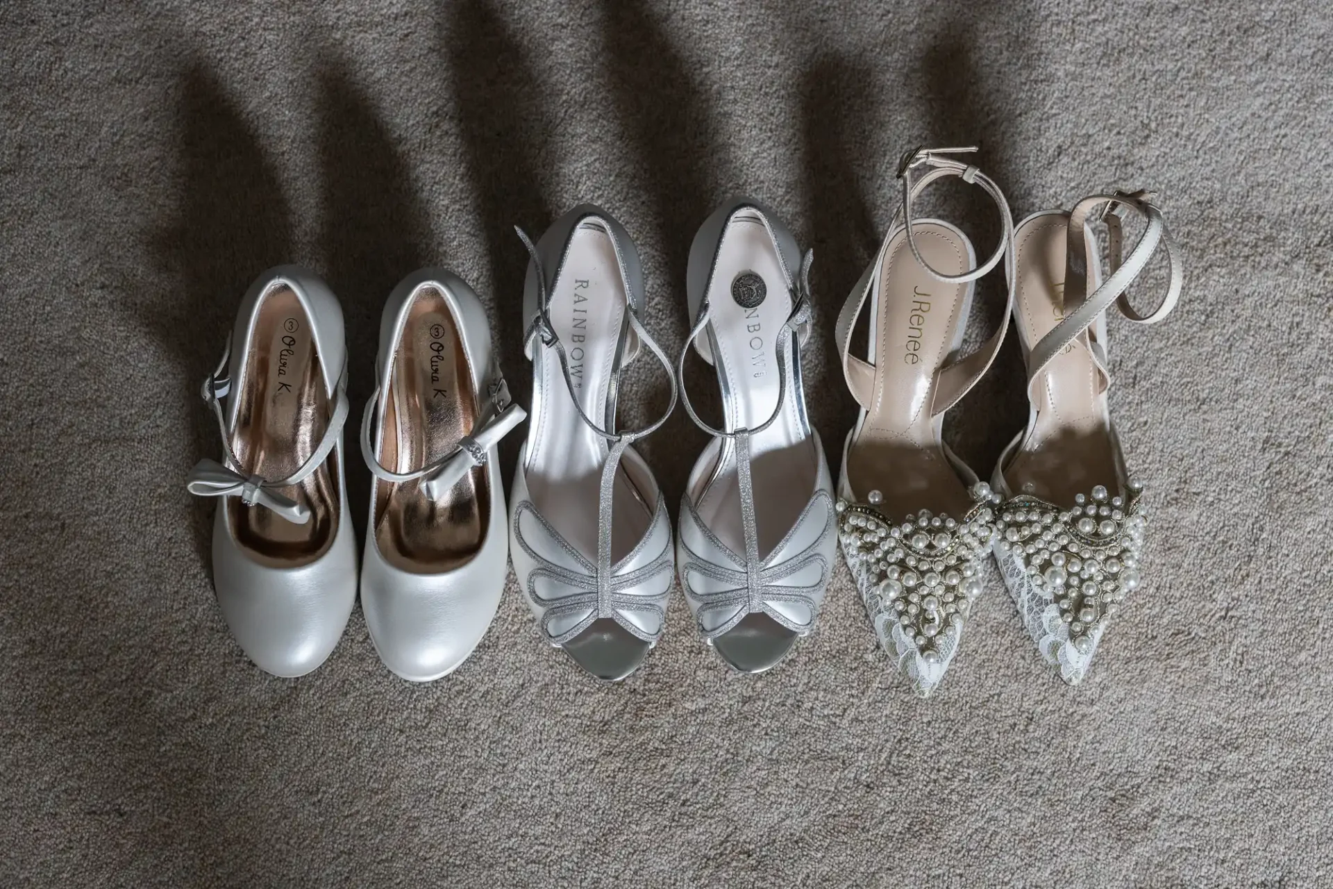 Three pairs of elegant women's shoes on a gray carpet: one with straps, one with open toes, and one with pearl embellishments.