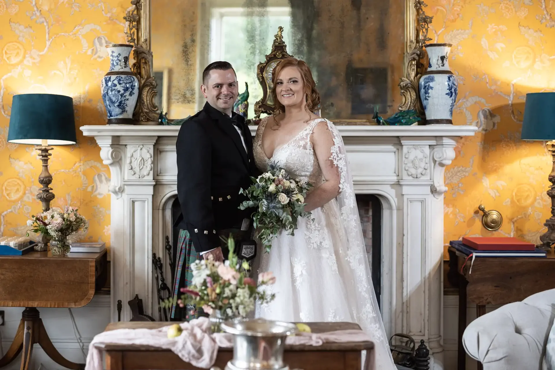 A couple in wedding attire stands in front of a fireplace with an ornate mirror, surrounded by vases and floral decorations, smiling at the camera.