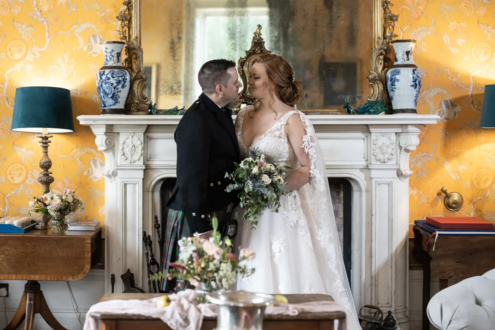 A couple in wedding attire stand in front of a fireplace. The room has a yellow floral wallpaper, blue vases, and a large mirror. A bouquet of flowers is in the foreground.