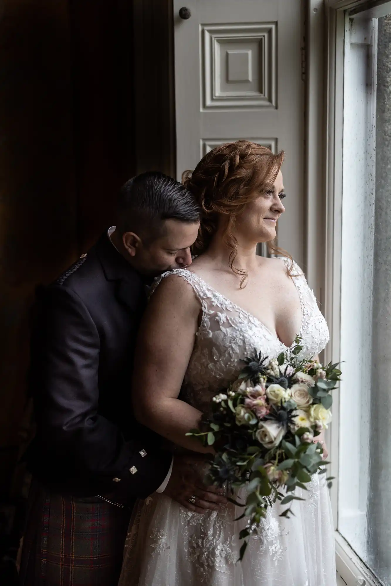 Wardhill Castle wedding photos: A couple stands by a window. The man embraces the woman, who holds a bouquet and wears a white wedding dress.