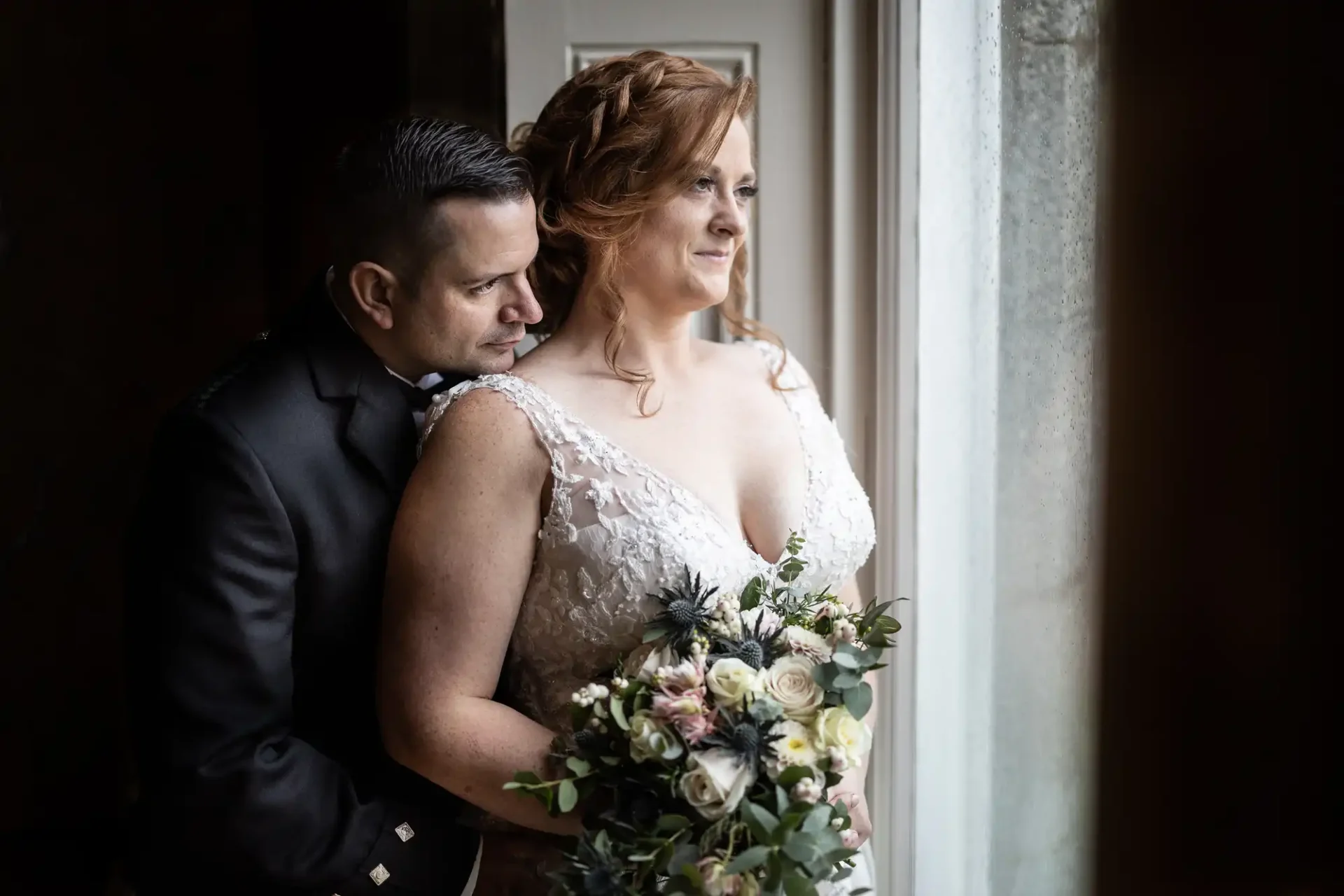 A couple stands close together, looking out a window. The woman holds a bouquet of flowers, and both are in formal attire.