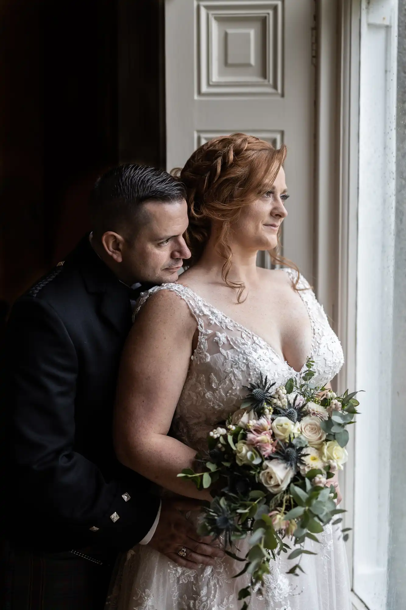 Wardhill Castle wedding photos: A bride in a lace gown holds a bouquet, standing by a window with a groom in a black suit embracing her from behind.