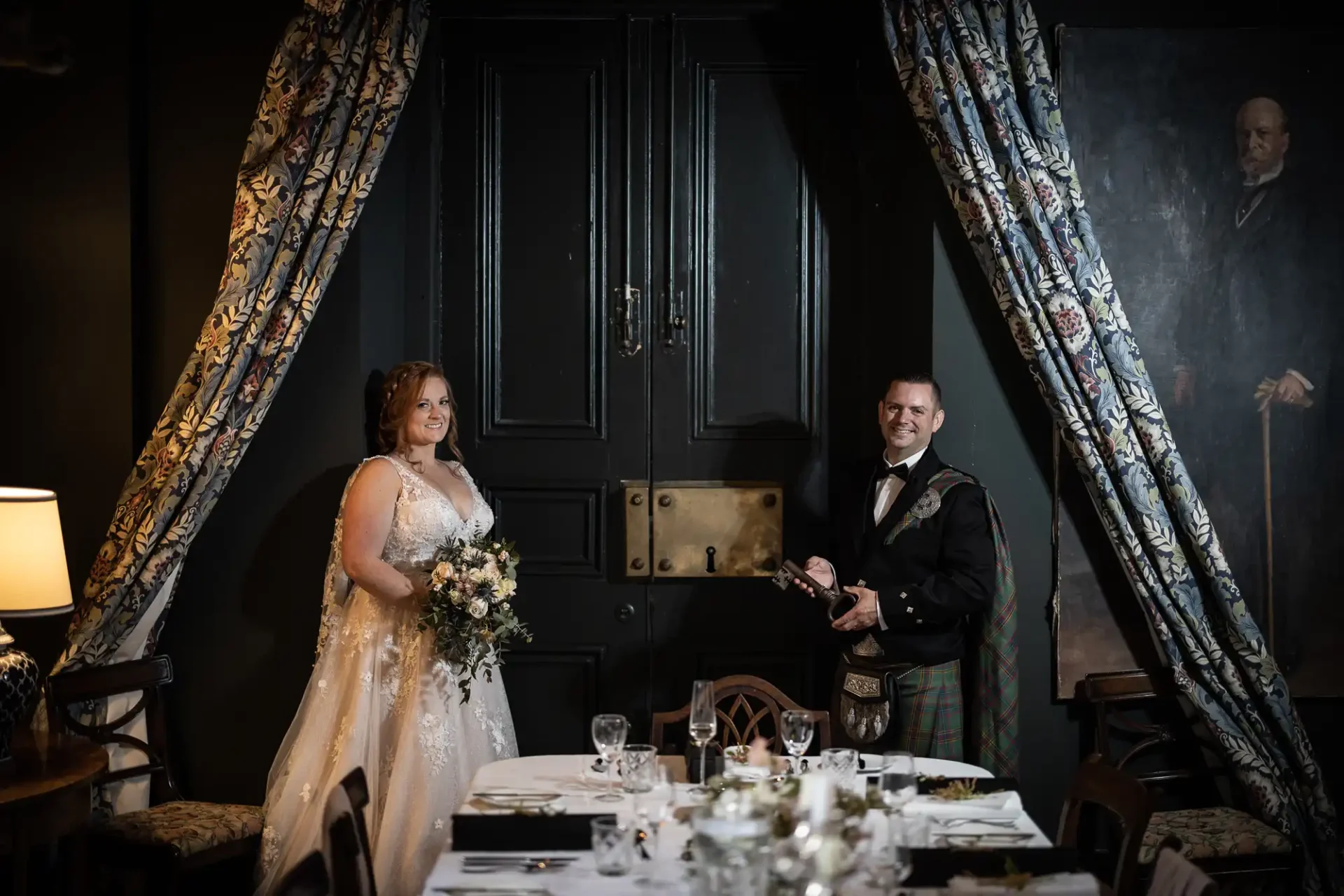 Wardhill Castle wedding photos: A bride and groom stand smiling in a dimly lit room with floral curtains and a large black door. The bride holds a bouquet; the groom is dressed in formal attire with a kilt.