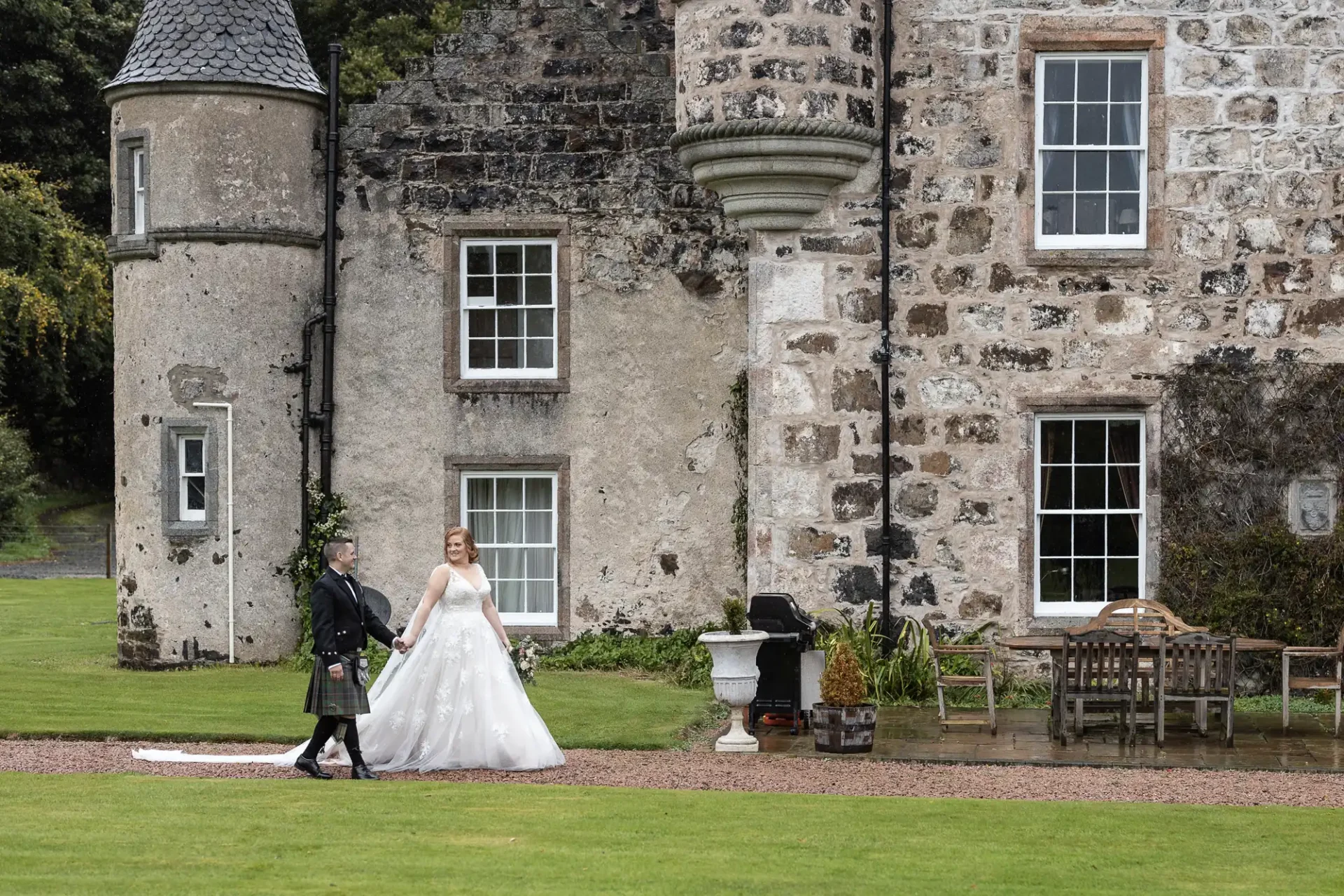 A bride and groom walk hand-in-hand on a lawn, near a large stone building with turrets and multiple windows. The groom is in traditional attire, and the bride wears a white gown.