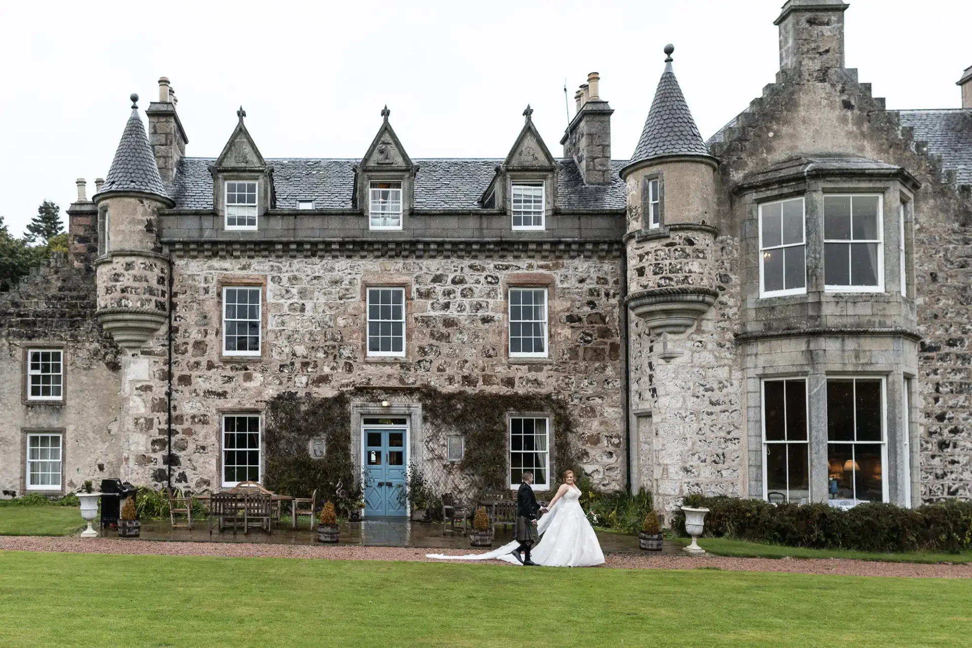 A bride and groom walk in front of a large, historic stone building with turrets and multiple windows, set on a grassy lawn.