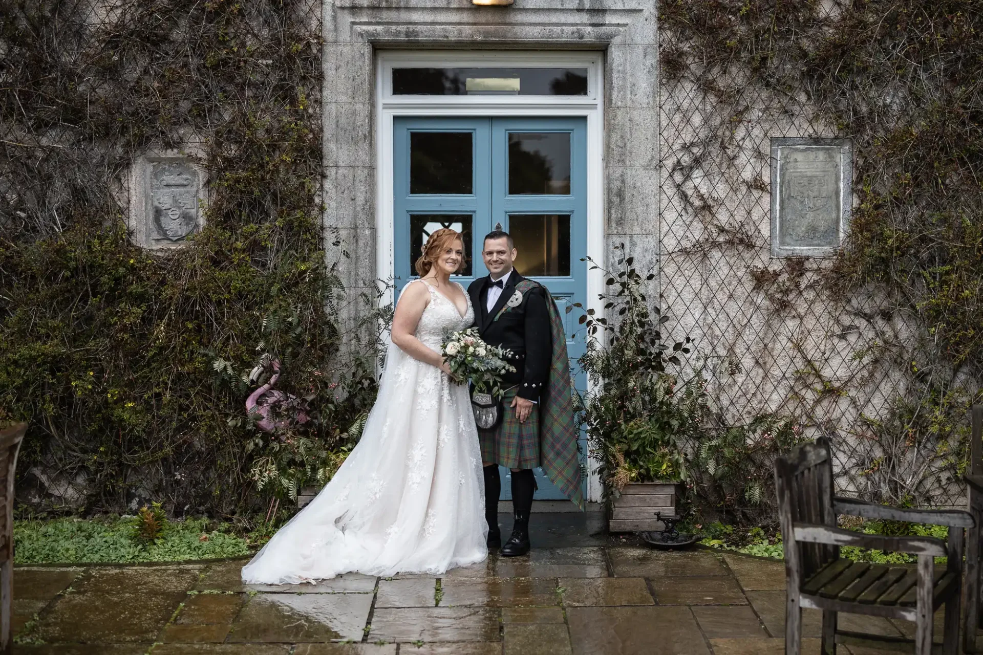 A bride and groom stand smiling in front of a vine-covered stone building entrance. The bride wears a white gown, and the groom wears traditional Scottish attire with a kilt.