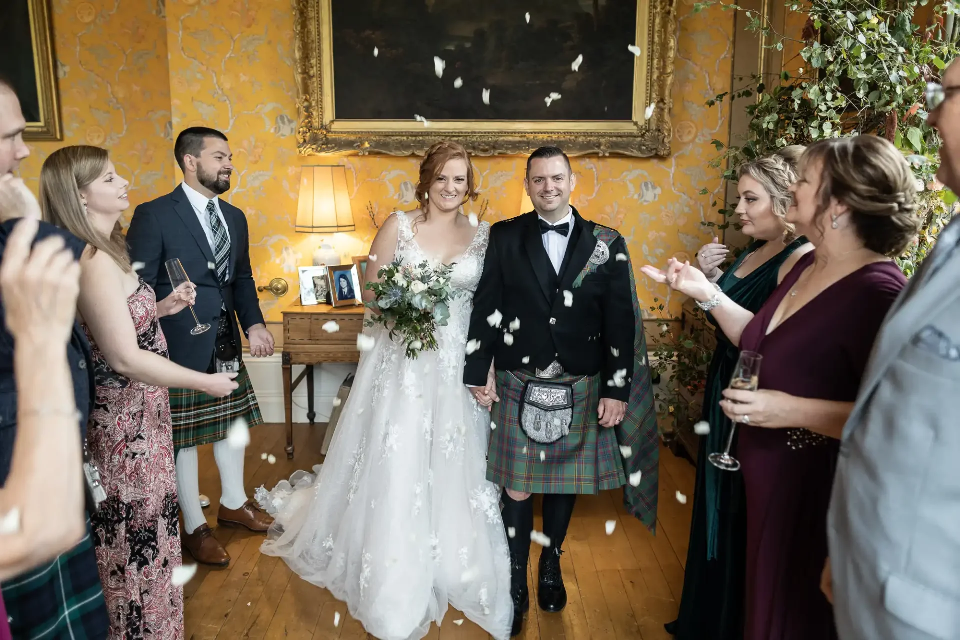 A bride and groom, with the groom in a kilt, smile as guests throw confetti around them in a warmly decorated room.