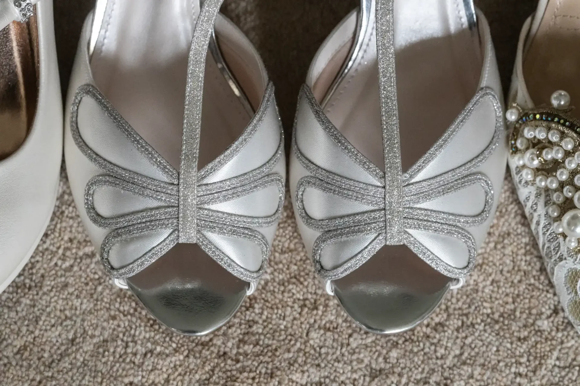 Silver strappy women's sandals with a butterfly-like design on the straps, placed on a textured beige carpet.