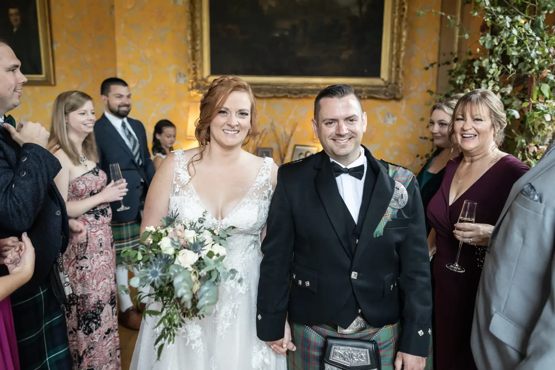 Bride and groom pose for a photo at their wedding, surrounded by smiling guests. The bride holds a bouquet, and the groom wears a formal outfit with tartan accents.