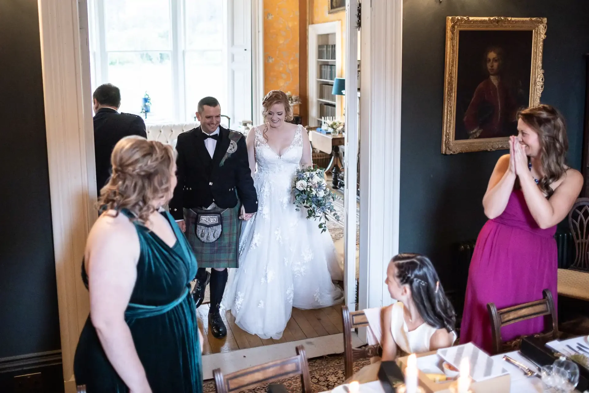 A couple in wedding attire enters a room as guests look on, smiling. The groom wears a kilt, and the bride holds a bouquet.