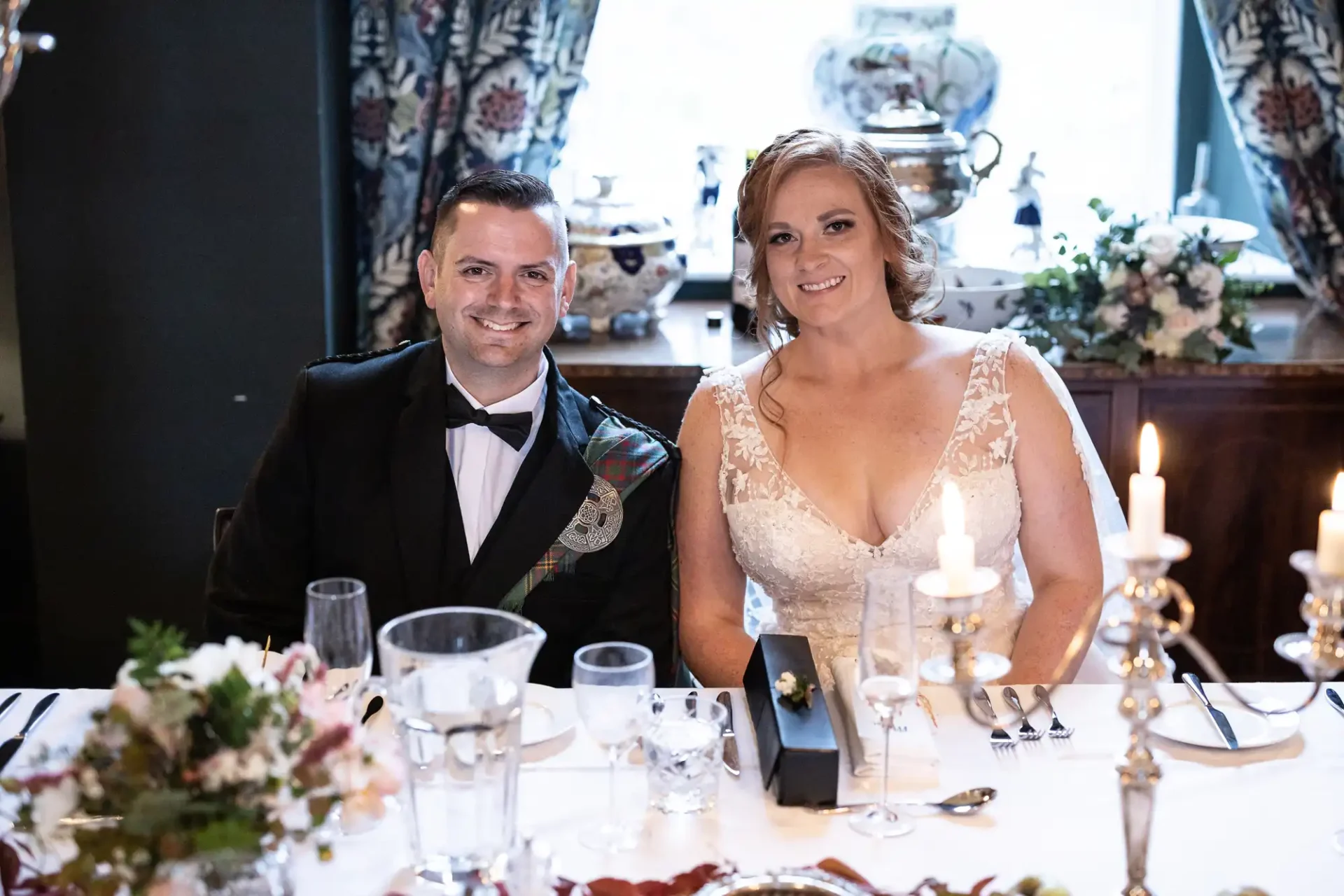 A couple in formal attire sits at a dining table decorated with flowers and candles, smiling for the camera.