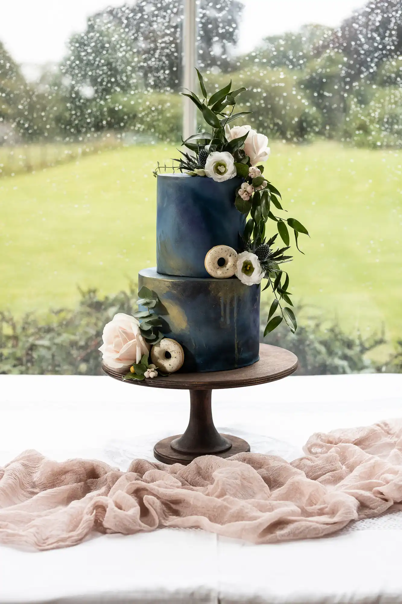 A two-tier dark blue cake with white flowers and greenery decor, placed on a wooden stand, with a beige cloth draped on the table in front of a rainy window overlooking a garden.