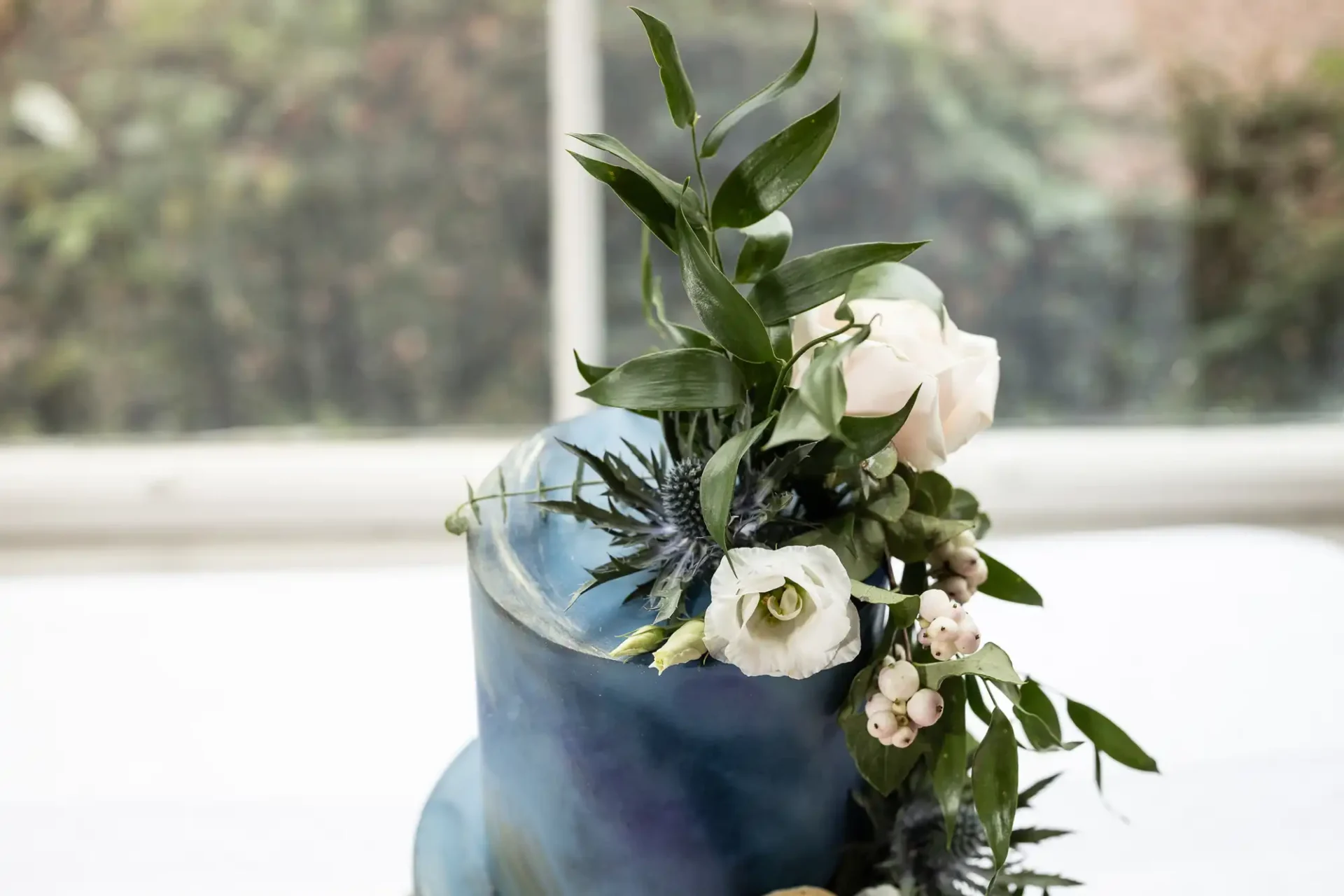 A blue ombre cake with white roses, greenery, and small white berries on top. The background is blurred.