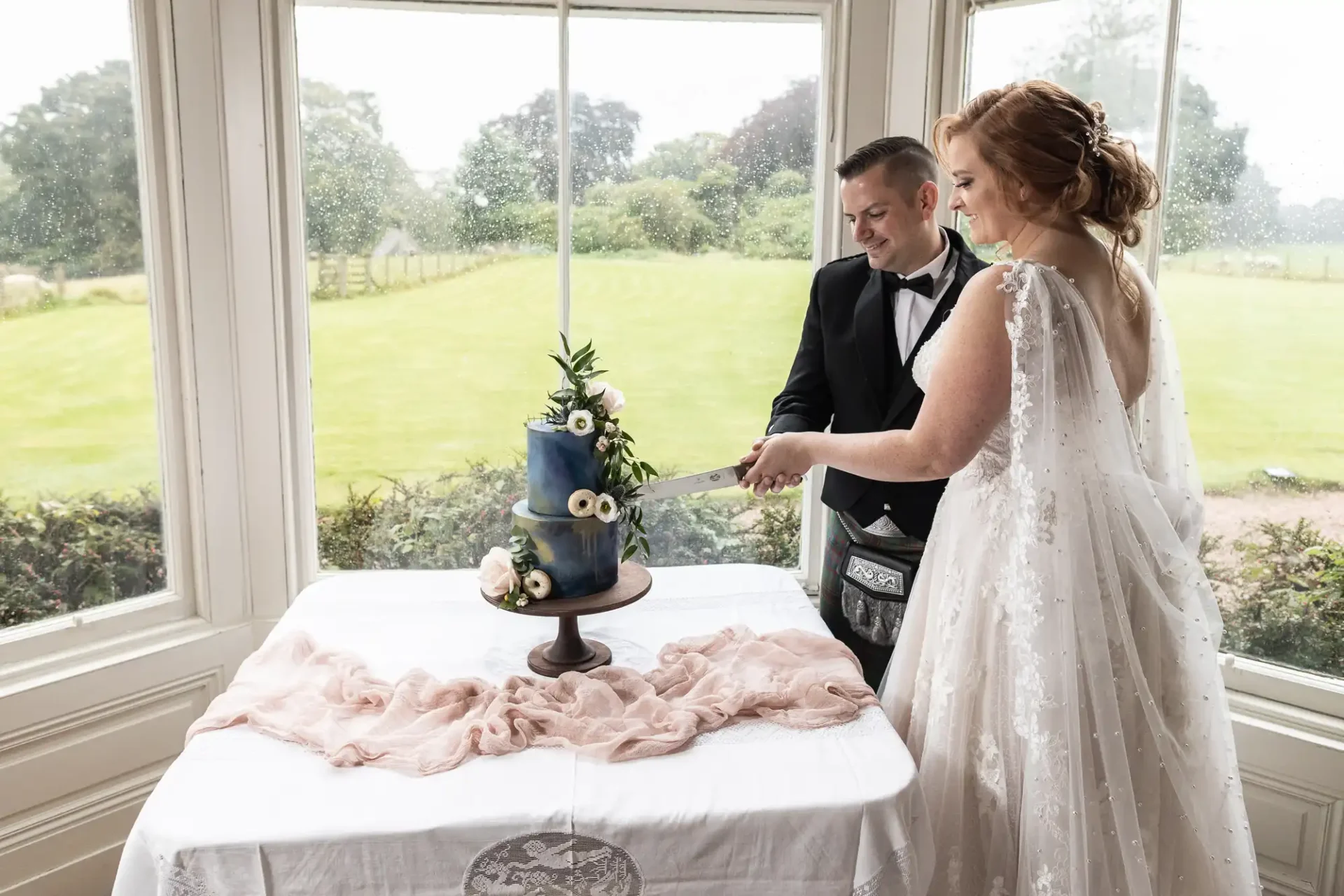 A bride and groom cut a blue tiered wedding cake adorned with flowers and blueberries, in front of a large window showing a garden view.