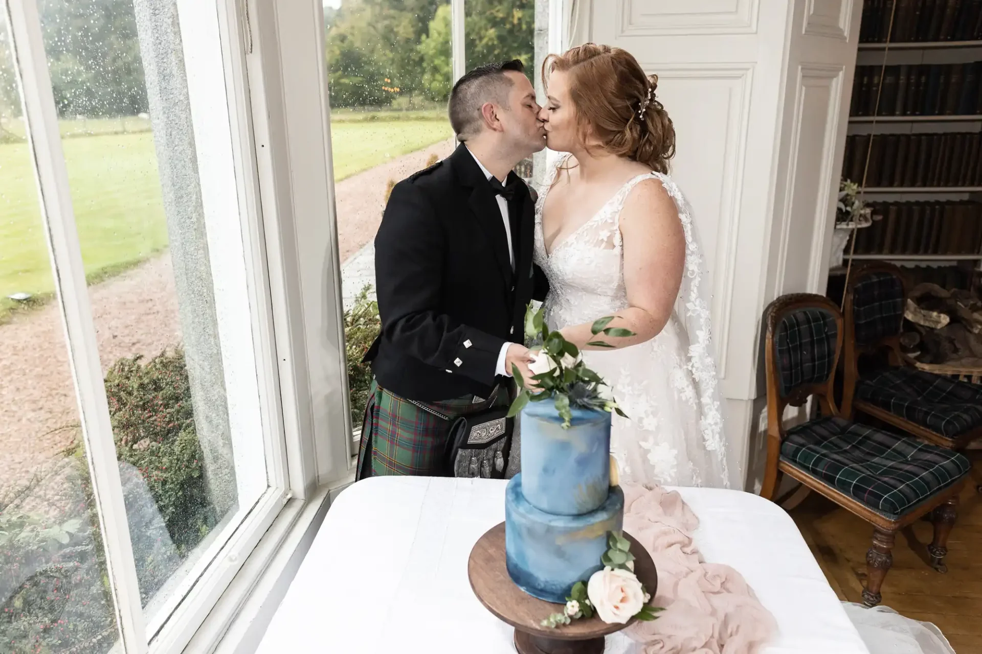 A couple in wedding attire kisses by a window. A blue cake with floral decorations is on a table in front of them.