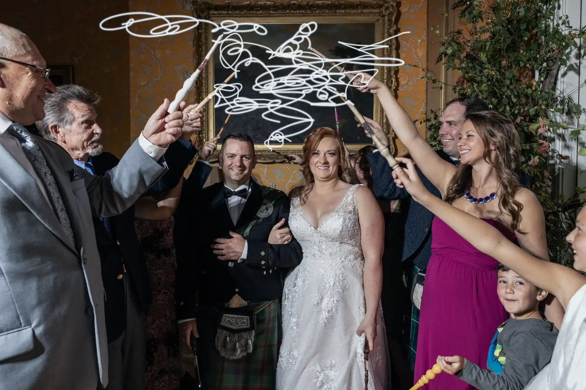 Bride and groom in formal attire smile as guests hold sparklers indoors, with a framed picture on the wall behind them.