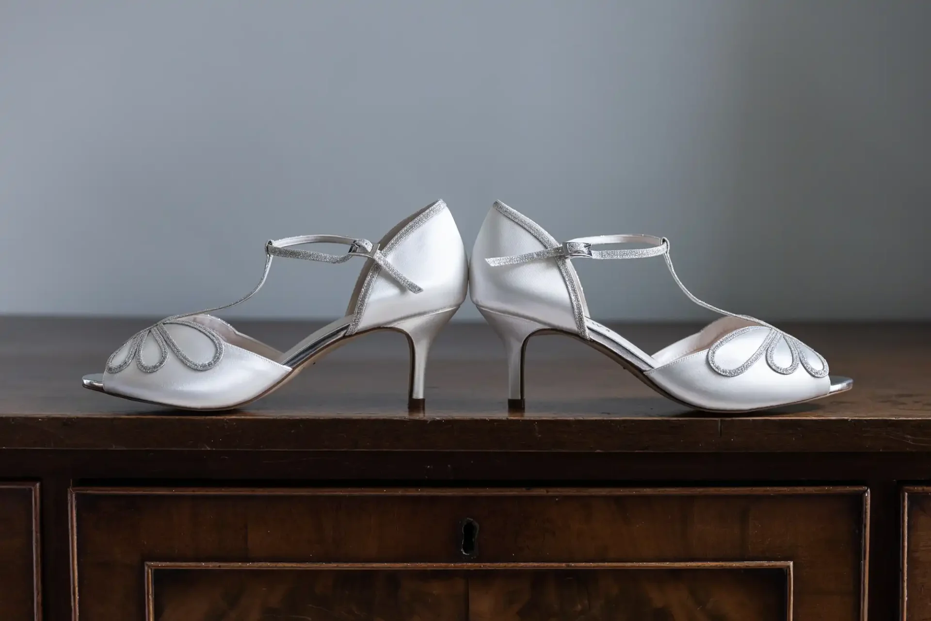 A pair of elegant white heeled sandals with ankle straps and decorative stitching sit on a wooden surface.