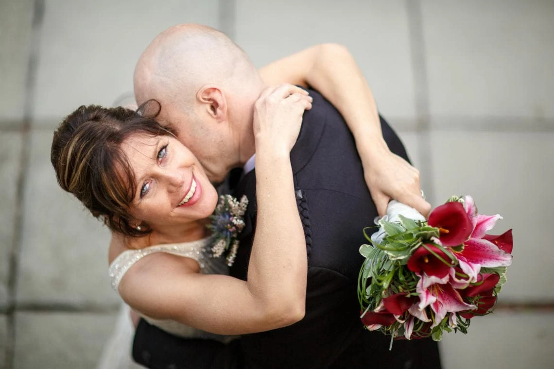 bride looks up at the camera as groom kisses her neck on the patio