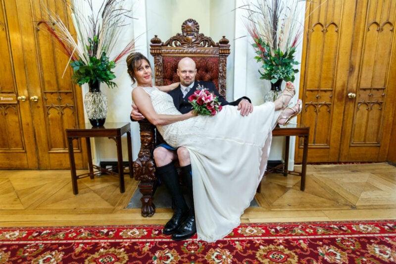 wedding photographer at Dalhousie Castle - bride sits on groom's lap on the throne in the reception area