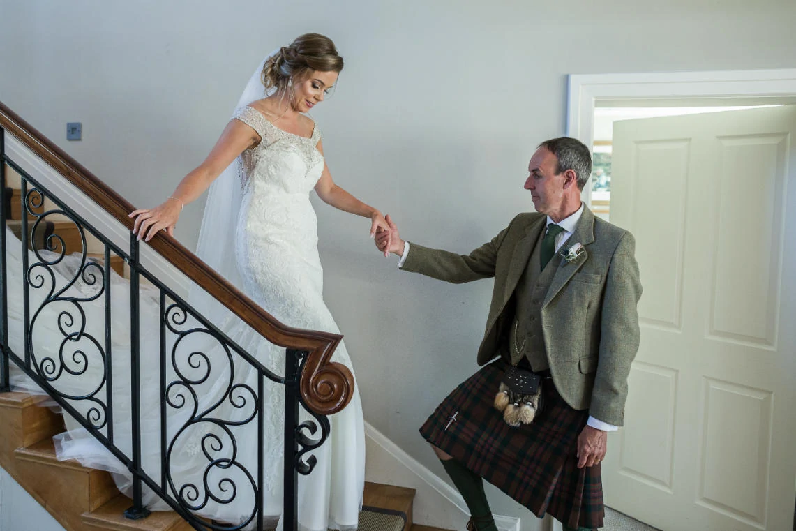 father of the bride helping the bride come down the stairs in her wedding dress