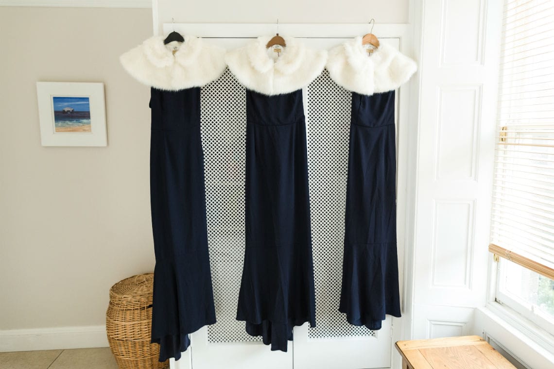 Three dark blue bridesmaids dress with white shrugs hanging up against white wardrobe