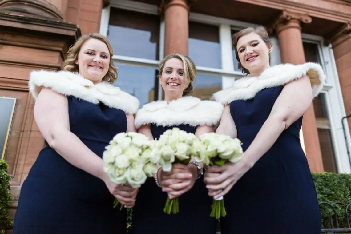 Three bridesmaids wearing dark blue dresses and white shrugs holding bouquets of flowers