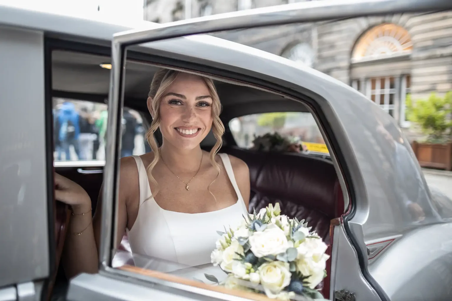 A woman in a white dress holding a bouquet of flowers sits in the back seat of a classic car with the door open, smiling at the camera.