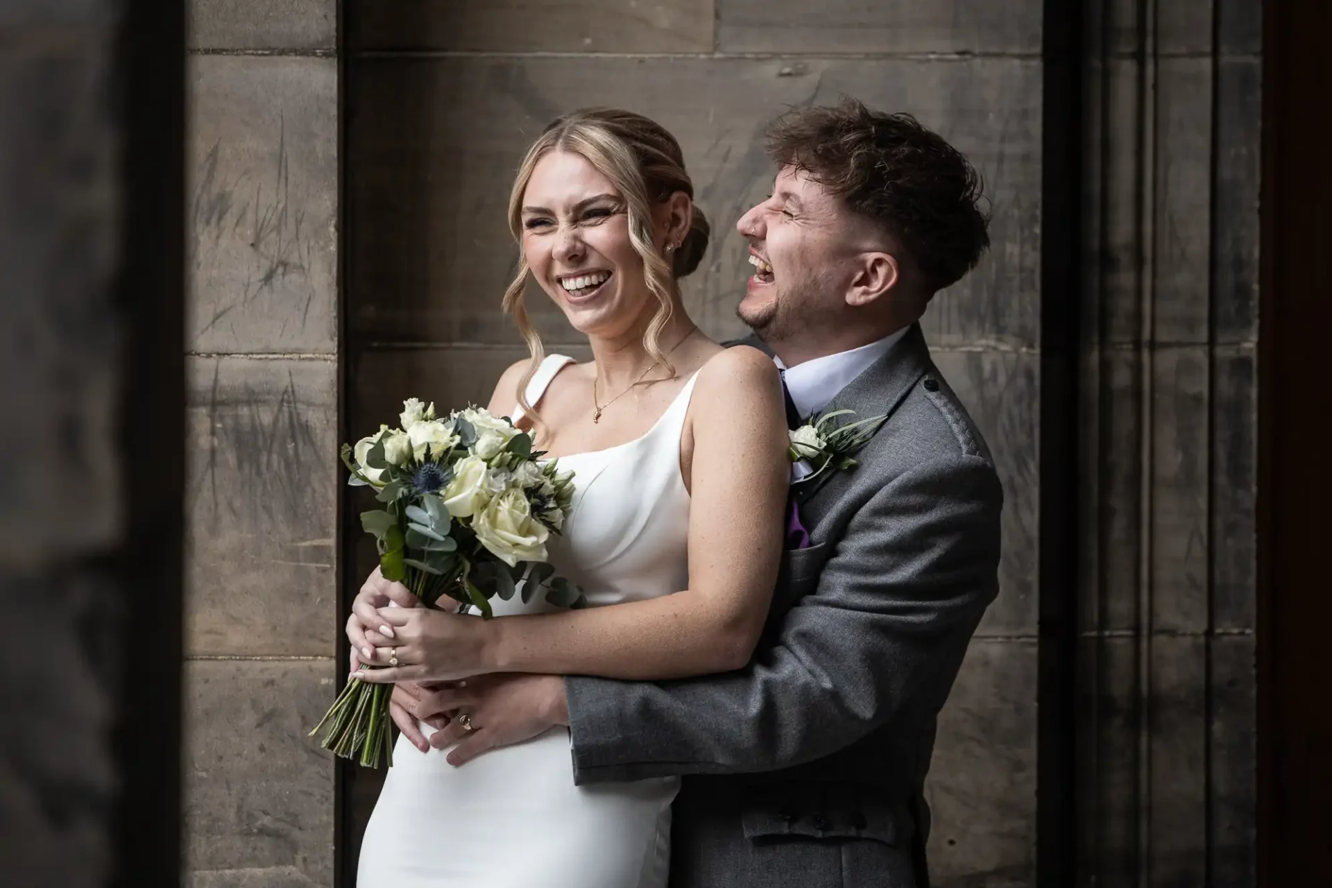 A bride in a white dress holding a bouquet and a groom in a grey suit laugh together while standing in front of a stone wall.