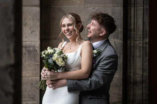 A bride in a white dress holding a bouquet and a groom in a grey suit laugh together while standing in front of a stone wall.