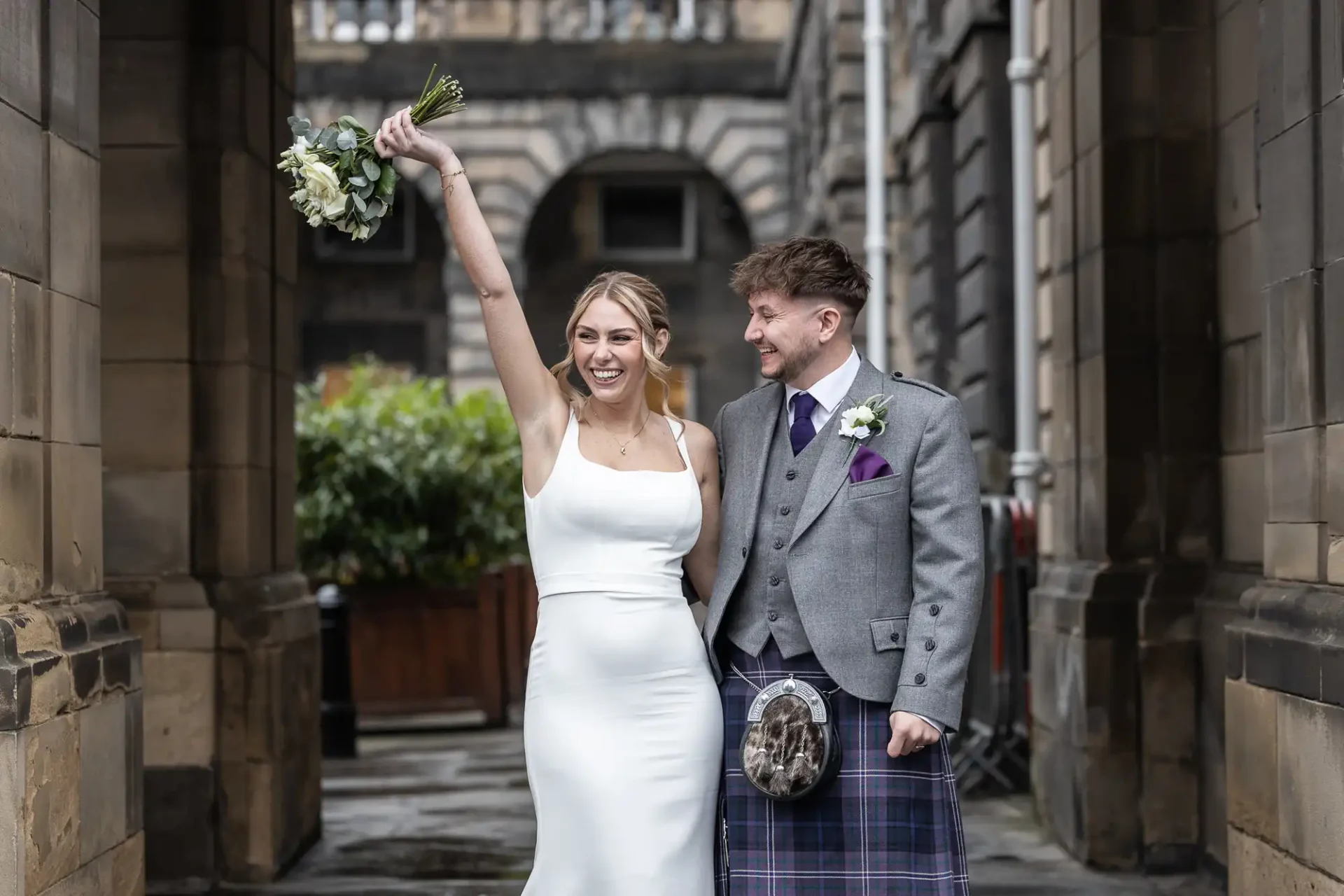 A bride in a white dress raises her bouquet joyfully at her elopement in Edinburgh, while standing next to a groom in a grey jacket and tartan kilt on a stone walkway.