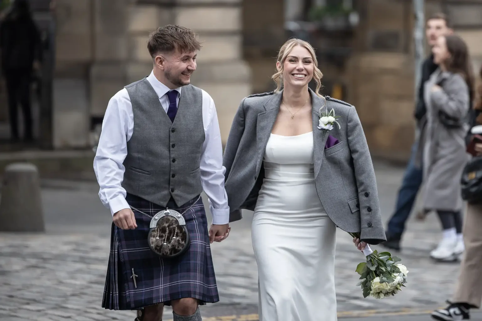 A smiling bride and groom walk hand in hand on a cobblestone street. The groom wears a kilt and vest; the bride wears a white dress and jacket, holding a bouquet.
